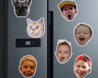 Könnte beinhalten: Eine Sammlung von Magneten in Gesichtsform, darunter eine französische Bulldogge, eine Katze und mehrere Kinder. Die Kinder tragen Hüte und machen verschiedene Gesichtsausdrücke. Die Magnete sind weiß mit weißem Rand und an einem grauen Kühlschrank befestigt.