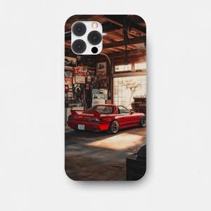 Op de afbeelding: Telefoonhoesje met een rode sportwagen in een garage. De auto heeft een achterspoiler en aangepaste wielen. De garage heeft een houten plafond en is gevuld met gereedschap. De scène wordt verlicht door warm zonlicht.