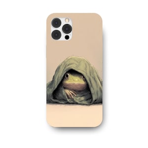 以下が含まれることがあります： 緑色の布の下から覗くカエルのイラストが描かれた携帯電話ケース。カエルは黄色い目と薄茶色の顔をしています。ケースはベージュ色の背景です。