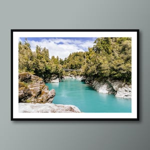 Puede incluir: Impresión enmarcada de un paisaje escénico. Un río turquesa fluye a través de un desfiladero rocoso, flanqueado por árboles verdes. Un pequeño puente colgante cruza el agua. El cielo es azul con nubes dispersas.