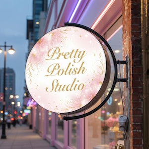 May include: A round, double-sided sign for a nail salon. The sign features the text "Pretty Polish Studio" in gold glitter lettering, surrounded by pink floral and leaf designs. The sign is mounted on a black metal frame.