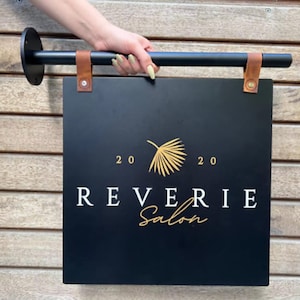 Może przedstawiać: Czarny prostokątny szyld salonu z napisem "REVERIE Salon" w białych i złotych literach. Znak jest trzymany przez czarny metalowy pręt z brązowymi sk&oacute;rzanymi paskami. Znak ma r&oacute;wnież złoty wz&oacute;r liścia i liczby "20 20".