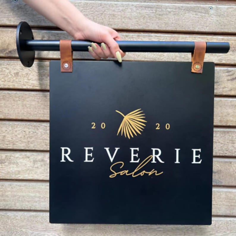 Może przedstawiać: Czarny prostokątny szyld salonu z napisem "REVERIE Salon" w białych i złotych literach. Znak jest trzymany przez czarny metalowy pręt z brązowymi sk&oacute;rzanymi paskami. Znak ma r&oacute;wnież złoty wz&oacute;r liścia i liczby "20 20".