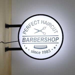 Puede incluir: Un letrero de barbería redondo e iluminado con el texto "PERFECT HAIRCUT BARBERSHOP since 1985". El letrero presenta un borde negro, un par de tijeras, un peine y estrellas. Está montado en una pared con soportes metálicos negros.