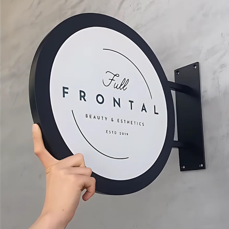 Może przedstawiać: Okrągły, dwustronny szyld z czarną ramą i białym tłem. Tekst "Full Frontal BEAUTY & ESTHETICS ESTD 2018" jest czarny. Zamontowany na czarnym metalowym wsporniku.