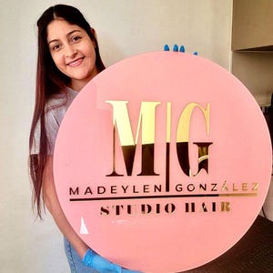 Könnte beinhalten: Ein rundes, rosa Schild mit goldfarbenen Buchstaben. Auf dem Schild steht "MG MADEYLEN GONZÁLEZ STUDIO HAIR." Das Schild wird von einer Person gehalten. Das Schild ist für einen Friseursalon.