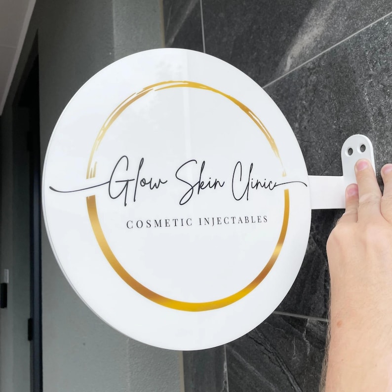 Może przedstawiać: Okrągły, biały szyld ze złotymi akcentami i napisem "Glow Skin Clinic" czarnym skryptem. Słowa "Cosmetic Injectables" znajdują się poniżej. Szyld jest zamontowany na białym wsporniku.