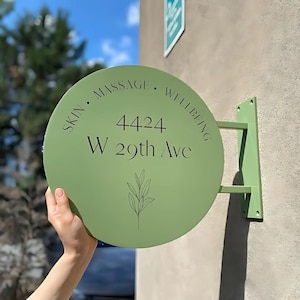 May include: A circular, pale green sign displaying the words "SKIN • MASSAGE • WELLBEING" in a curved layout. The address "4424 W 29th Ave" is centered, with a botanical illustration beneath. The sign is mounted on a green bracket.