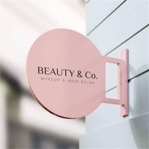 Puede incluir: Un letrero rosa y circular para "BEAUTY & Co. Makeup & Hair Salon" está montado en una pared. El letrero es de color rosa sólido con el nombre de la empresa y el servicio en texto negro. El letrero está sujeto a la pared con un soporte rosa a juego.