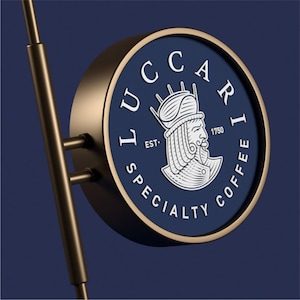 Może przedstawiać: Okrągły, niebieski i złoty znak z tekstem "LUCCARI EST. 1750 SPECIALTY COFFEE" i białą ilustracją osoby w koronie.