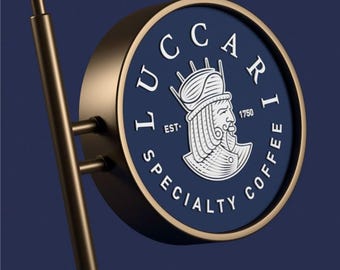 Könnte beinhalten: Ein rundes, blaues und goldenes Schild mit dem Text "LUCCARI EST. 1750 SPECIALTY COFFEE" und einer weißen Illustration einer Person mit einer Krone.