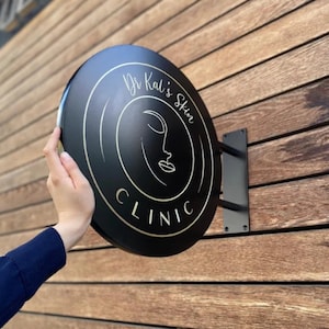 Może przedstawiać: Czarny, okrągły szyld dla "Dr Kal's Skin Clinic" ze złotymi literami i minimalistycznym wzorem twarzy. Szyld jest zamontowany na drewnianej ścianie z czarnym metalowym wspornikiem.