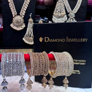 Könnte beinhalten: Eine Auswahl an aufwendigem Schmuck, darunter Halsketten, Ohrringe und Armreifen. Die Armreifen sind silber- und goldfarben, mit glitzernden Steinen und hängenden Anhängern verziert. Die Halsketten und Ohrringe werden auf schwarzen Ständern präsentiert.