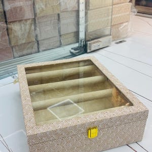 Op de afbeelding: Een rechthoekige sieradendoos met een beige textuur aan de buitenkant en een herhalend geweven patroon. De doos heeft een doorzichtig glazen deksel en een goudkleurige metalen sluiting. Binnenin bevinden zich vijf rijen voor ringen.