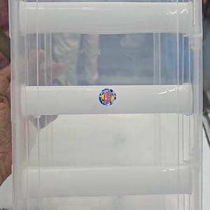 May include: A clear plastic storage container with a rectangular shape. The container has a lid and several white horizontal supports. A small, round sticker with blue, white, and gold accents is visible on the front. The container is designed for storage.