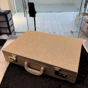 May include: A rectangular, gold-coloured briefcase with a textured pattern. It features a handle, two combination locks, and gold-coloured hardware. The briefcase is resting on a black and white surface.