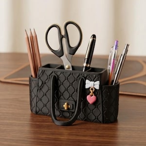 May include: A black desk organizer shaped like a handbag with a quilted texture, featuring a gold-tone clasp, a white bow charm, and a pink heart charm. The organizer holds three wooden pencils, a pair of black scissors, and three pens.