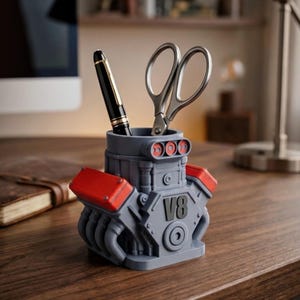 May include: A grey V8 engine block shaped desk organiser featuring red valve covers and black accents. The organiser holds a black fountain pen and a pair of silver metal scissors. The front of the engine block is clearly embossed with the text V8.