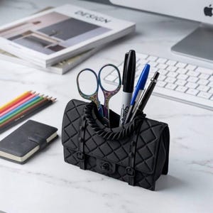 May include: A black, quilted-texture desk organizer shaped like a small handbag. It features a braided handle, two vertical straps with buckles, and a central clasp. Inside are a pair of iridescent scissors, a black marker, and two pens.