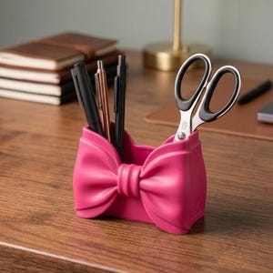 May include: A bright pink desk organizer shaped like a bow, holding three pens and a pair of scissors with black handles. The organizer has a smooth, molded surface and a central knot detail, providing a functional storage space for office supplies.