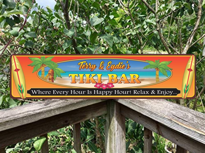 The tiki bar sign is mounted on a wooden deck railing surrounded by lush green foliage, giving it a relaxed outdoor tropical vibe that highlights its long horizontal shape.