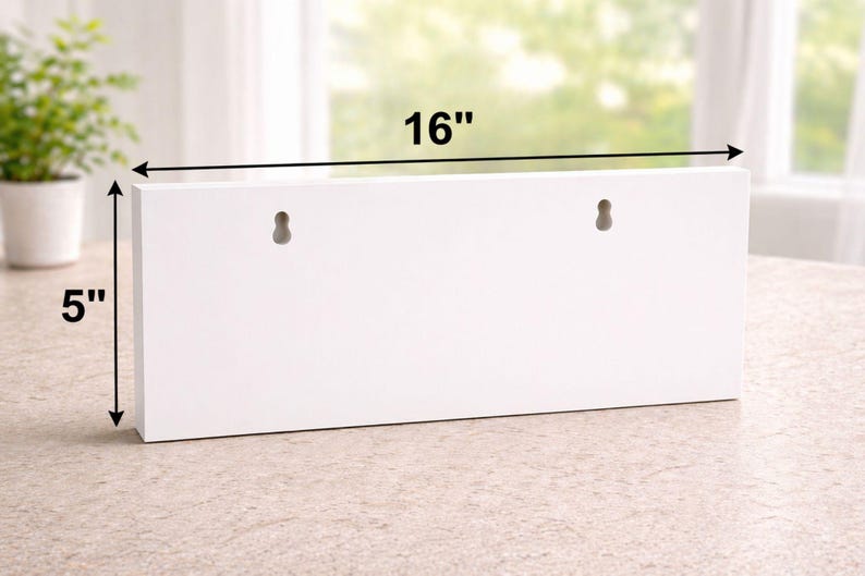 May include: A white rectangular wooden shelf, 40.6 cm long and 12.7 cm high, with two keyhole slots for hanging. The shelf is displayed on a light-colored surface, with a plant in a white pot visible in the background.