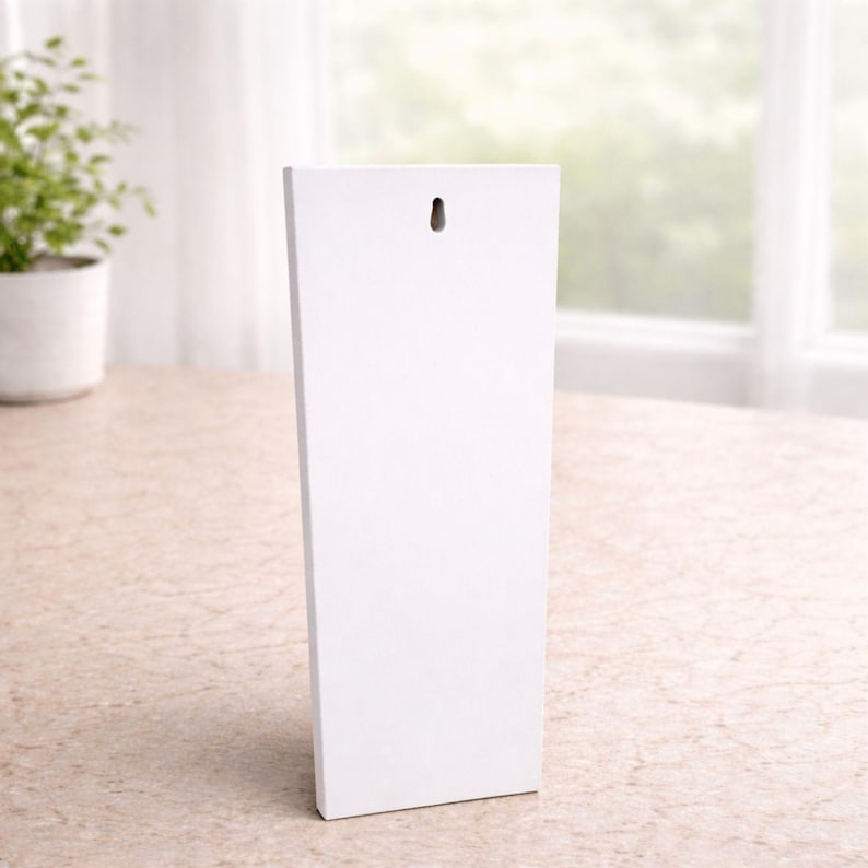 May include: A white rectangular wooden wall hanging with a hole at the top for hanging. The wall decor is leaning slightly to the left. The background includes a plant in a white pot and a window.