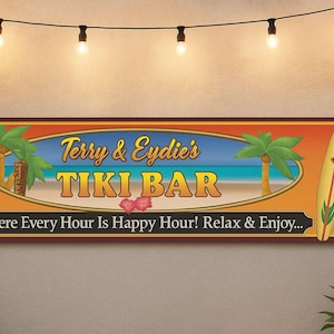 A rectangular tiki bar sign with a tropical beach scene and surfboards on each end is mounted on a light stucco patio wall. A string of warm Edison bulbs hangs above, and lush green plants frame the scene, creating a relaxed tropical ambiance.