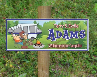 May include: A blue and green camping sign with a cartoon image of a couple sitting by a campfire in front of a camper. The sign reads "Terry & Eydie Adams Welcome to our Campsite!"