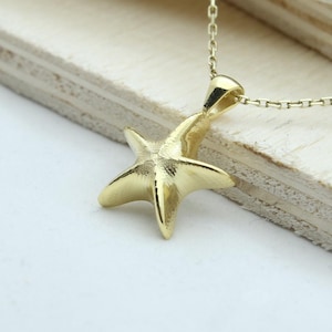 Pode incluir: Um colar com pingente de estrela do mar dourada. O pingente de estrela do mar é detalhado e suspenso por uma corrente delicada. O colar é exibido em uma superfície de madeira clara, com um fundo branco.