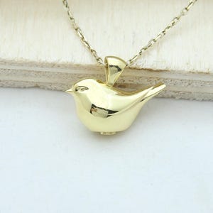 Könnte beinhalten: Eine goldfarbene Vogel-Anhänger-Halskette. Der Vogel ist ein stilisiertes, dreidimensionales Design mit glatter, polierter Oberfläche. Die Halskette ist zierlich und goldfarben. Der Hintergrund ist eine helle Oberfläche mit Holzbrettern.