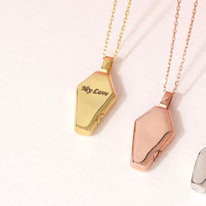 Puede incluir: Tres collares de oro, oro rosa y plata con colgantes en forma de ataúd. El colgante de oro tiene la inscripción "My Love".