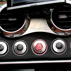Può includere: Primo piano del cruscotto di un'auto con bocchette dell'aria con finiture cromate e pulsanti di controllo. La console centrale ha una placca argentata con la scritta "TT Roadster". Pulsanti: sedile riscaldato, luci di emergenza ed ESP.
