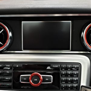Op de afbeelding: Close-up van een auto dashboard met een zwart infotainment scherm en zilveren afwerking. Het dashboard heeft radio-, navigatie- en telefoonbediening. Een rode draaiknop met het woord "ON" staat in het midden.