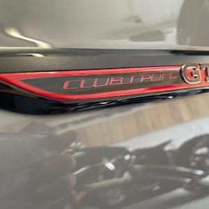 Peut inclure: Une voiture noire avec un emblème "Clubsport GTI" rouge et noir à l'arrière.