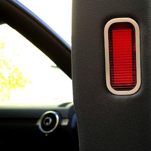 Op de afbeelding: Close-up van een auto-interieurdetail. Een rechthoekig rood licht met een zilveren frame is in een zwart, getextureerd paneel geplaatst. De binnenkant van de auto is zichtbaar op de achtergrond.