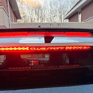 Könnte beinhalten: Nahaufnahme der Heckscheibe eines Autos mit einer roten LED-Lichtleiste. Die Lichtleiste hat ein Wabenmuster und den Schriftzug "CLUB SPORT" in Weiß. Die Heckscheibe ist schwarz.