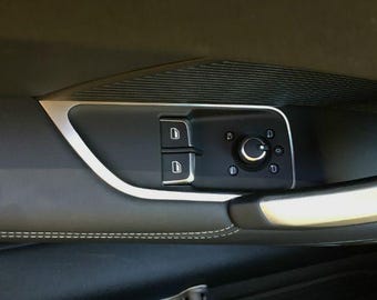 May include: Close-up of a car door panel featuring a black control panel with window and mirror adjustment buttons. The panel has a silver trim and is set against a dark gray interior with white stitching.
