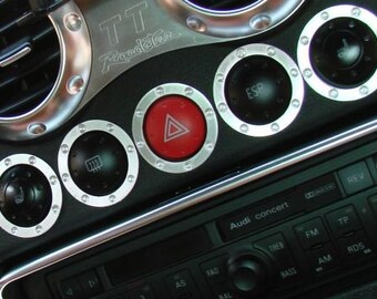 May include: Close-up of a car dashboard featuring silver trim and black controls. Includes air vents, circular buttons with silver trim, and a red hazard light button. The radio has the text "Audi concert".