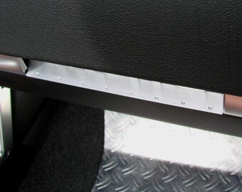 May include: Close-up of a vehicle interior detail. A black textured surface is above a silver metal trim piece with small rivets. Below, a diamond-plate metal surface is visible.