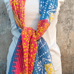 Puede incluir: Bufanda colorida con diseño de patchwork. Presenta secciones de tela rosa, amarilla y azul con costuras blancas. La bufanda está envuelta alrededor de un cuello alto blanco.