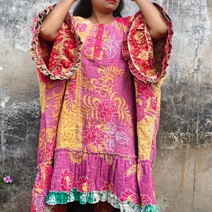 Könnte beinhalten: Eine gemusterte Tunika in Rosa, Gelb und Rot. Das Kleid hat ein Blumenmuster, Rüschenärmel und einen Stufenbund. Die Ärmel haben einen kontrastierenden roten und goldenen Stoff. Das Kleid wird mit schwarzen Leggings getragen.