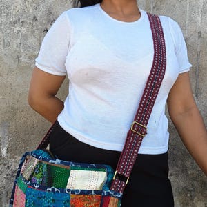 Pode incluir: Uma bolsa tiracolo com design de patchwork apresentando v&aacute;rios quadrados de tecido coloridos em tons de azul, rosa, laranja e branco, acentuada por bordados vis&iacute;veis de ponto corrido e uma al&ccedil;a longa ajust&aacute;vel com um padr&atilde;o vermelho escuro e azul.