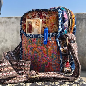 Può includere: Una borsa a tracolla multicolore con chiusura a zip e tracolla lunga. La borsa presenta vari tessuti nei toni del rosso, blu, arancione e rosa. Una custodia per telefono e un'etichetta blu con la scritta "Resuwas" sono visibili.