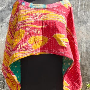 May include: A colourful, reversible, quilted top in red, yellow, and green, displayed on a black mannequin. The top features a floral pattern and is stitched with white thread. The neckline is trimmed with pink and blue fabric.