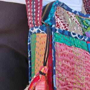 Pode incluir: Uma bolsa de ombro de patchwork com uma variedade de quadrados de tecido coloridos em tons de rosa, verde, azul e laranja. A bolsa &eacute; constru&iacute;da com costuras vis&iacute;veis em estilo kantha e inclui uma al&ccedil;a grossa tecida em bord&ocirc; e branco.