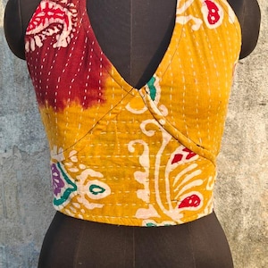 May include: A halter top with a patchwork design. The top features a V-neck and is made of fabric with a yellow and maroon color scheme. The fabric has a floral pattern with white, red, and green accents. The top is displayed on a black mannequin.