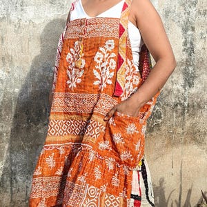 Pode incluir: Vestido avental laranja e branco com estampas florais e geométricas. O vestido tem bainha com babados e bolsos. A pessoa está usando um top branco por baixo. O vestido é feito de tecido acolchoado.