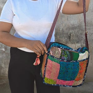 Pode incluir: Uma bolsa tiracolo retangular com um design de patchwork feito de v&aacute;rios quadrados de tecido coloridos em tons de rosa, azul, verde e laranja. A bolsa tem uma al&ccedil;a de ombro longa com padr&atilde;o escuro e uma pequena borla de tecido no z&iacute;per.