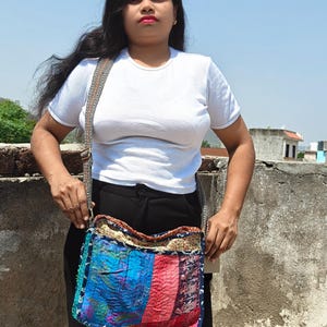 Pode incluir: Uma bolsa tiracolo retangular com um design de patchwork de pain&eacute;is de tecido de cores vibrantes em tons de azul, vermelho e amarelo. A bolsa possui uma al&ccedil;a cinza larga e texturizada, e um fecho de correr superior com costuras vis&iacute;veis.